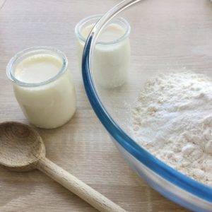 Plat contenant de la farine. 2 pots de yaourt à coté et une cuillère en bois